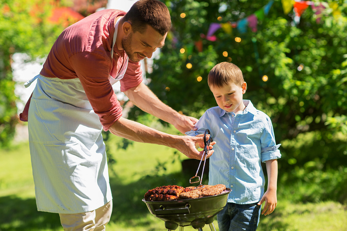 A father and son bbq meat together at a grill for father's day, outside, surrounded by trees and grass.
