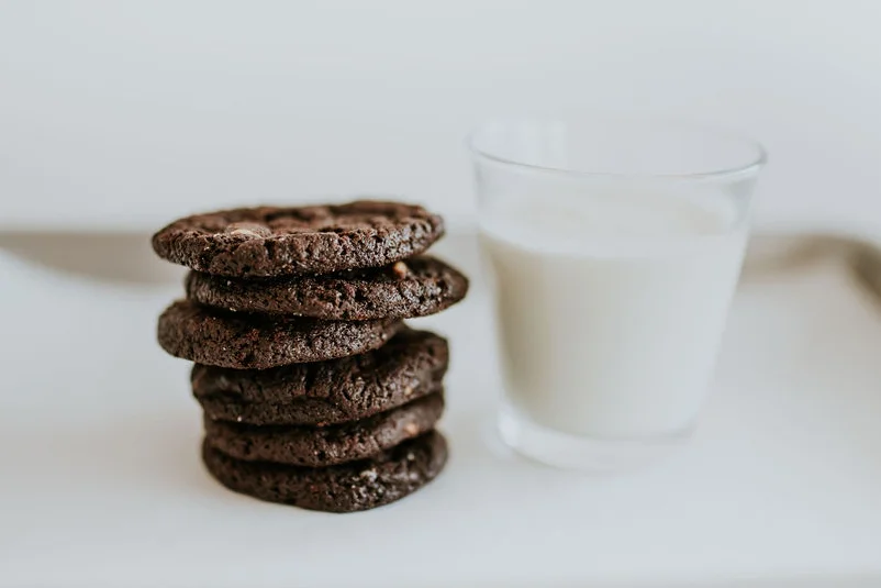 A stack of gourmet chocolate cookies on a white table next to a glass of milk - the perfect gift idea for chocolate lovers.